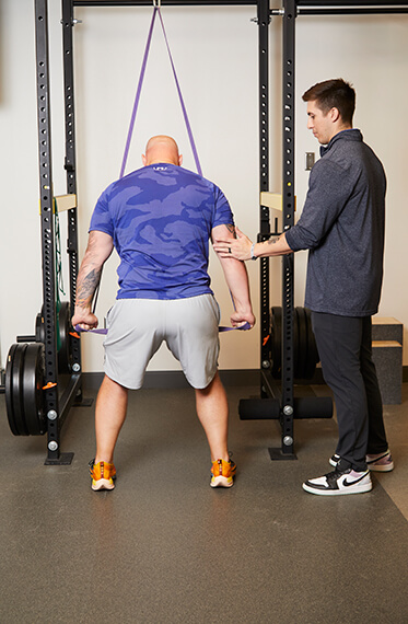 A patient facing away from the camera and using a band to upper body exercises while a therapist is beside him